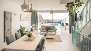 A modern living room with a dining area, a view of the pool, and a staircase leading to a balcony. - CALERO DREAM by Buenavilla | 3 bedroom  Frontline villa (Puerto Calero)