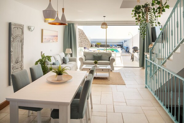 A modern living room with a dining area, a view of the pool, and a staircase leading to a balcony.