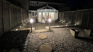 Ein überdachter Patio-Bereich mit einer Sitzgruppe und einer gläsernen Struktur, umgeben von einem Kiesweg und einem Holzzaun.