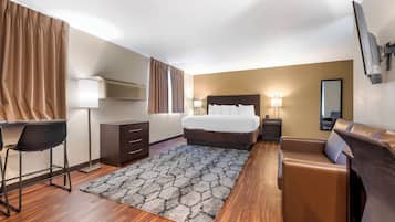 A hotel room with a bed, a desk with a chair, a sofa, a lamp, a mirror, and a patterned rug.