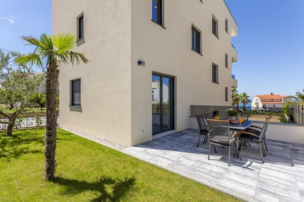 A modern two-story building with a patio area featuring a table and chairs, a palm tree, and a clear view of the surrounding neighborhood.