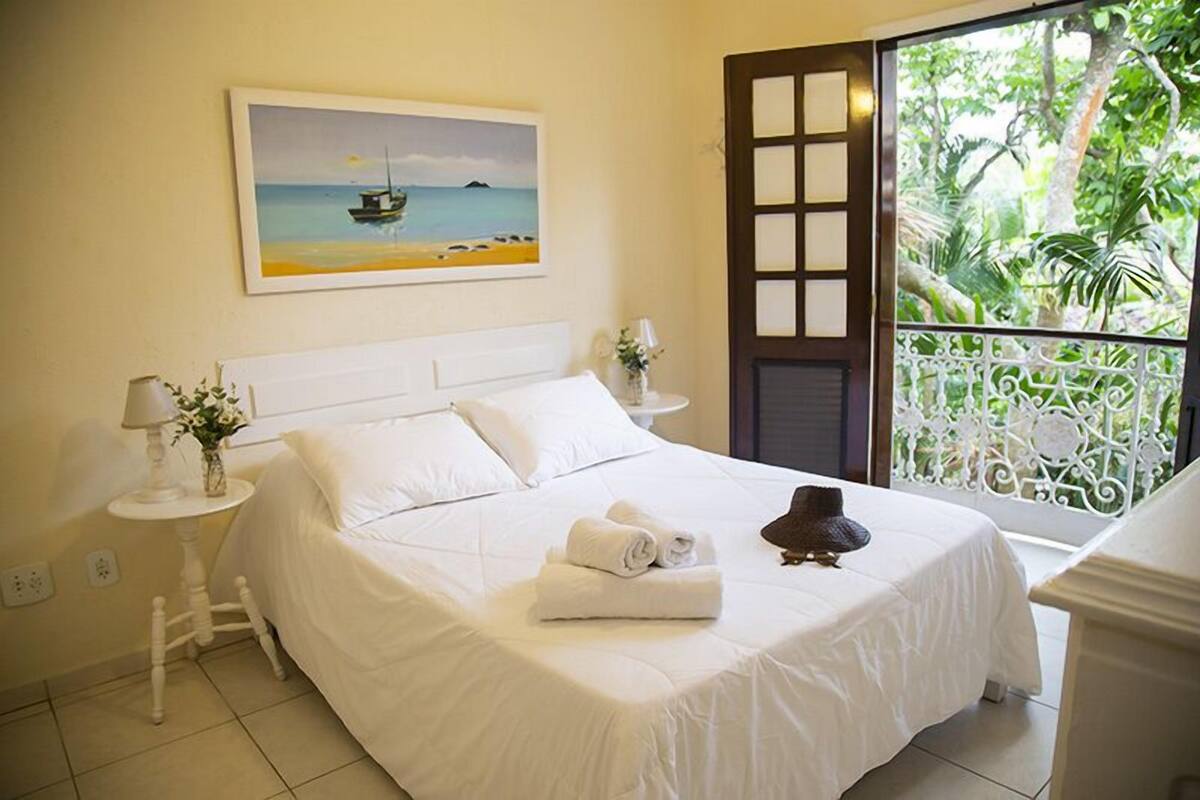 Ein ordentlich bezogenes Bett mit weißen Leinen, ein Gemälde mit Strandmotiv und ein Balkon mit schmiedeeisernem Geländer.