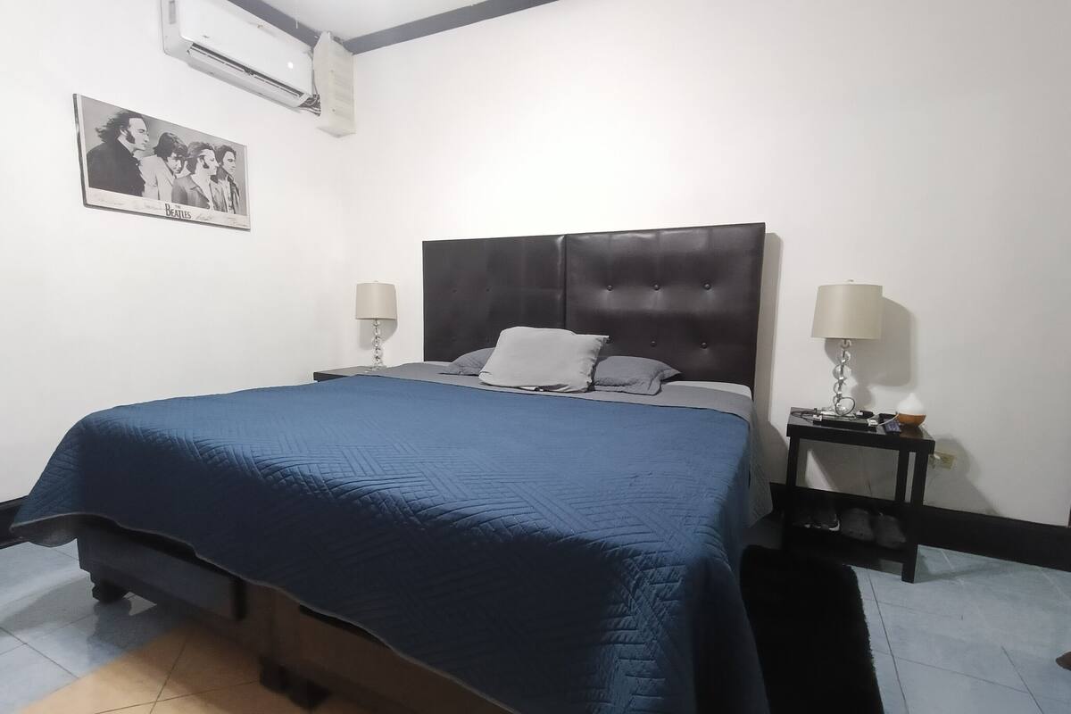Ein Schlafzimmer mit einem großen Bett, zwei Nachttischen mit Lampen, einer Wandklimaanlage und einem gerahmten Bild an der Wand.