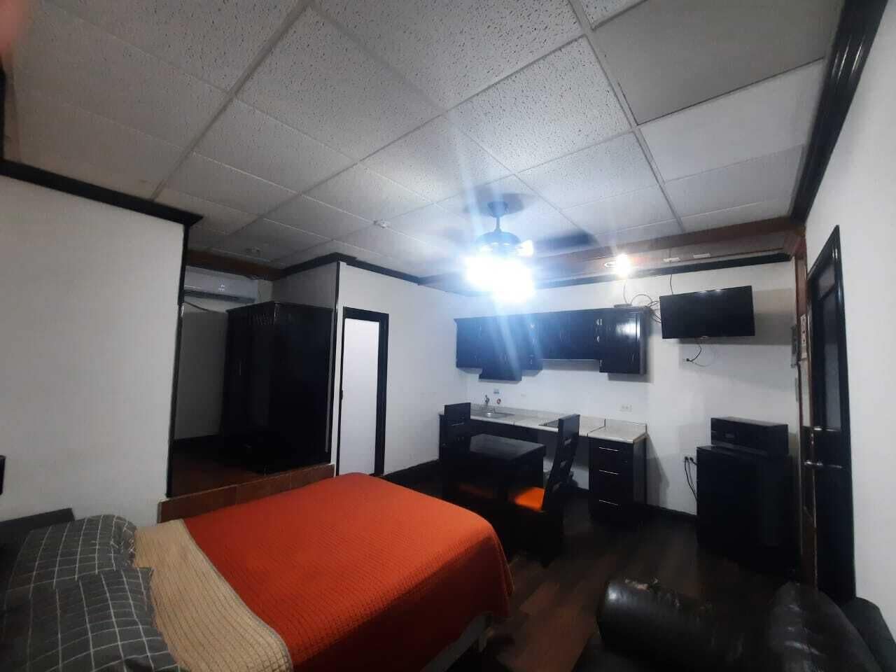 A hotel room with a bed, a desk with a chair, a television mounted on the wall, and a wardrobe.