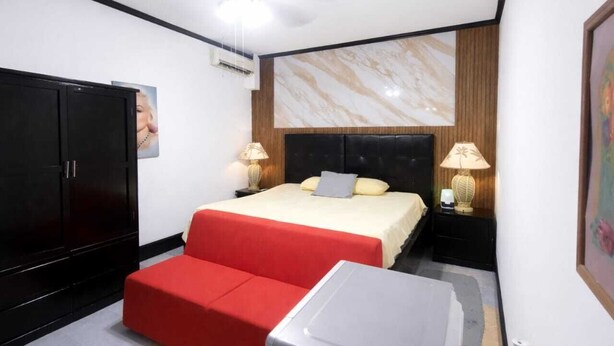 A hotel room with a bed, two bedside lamps, a red sofa, a small table, and a wardrobe. - Hotel Garcias Suites (Linares)