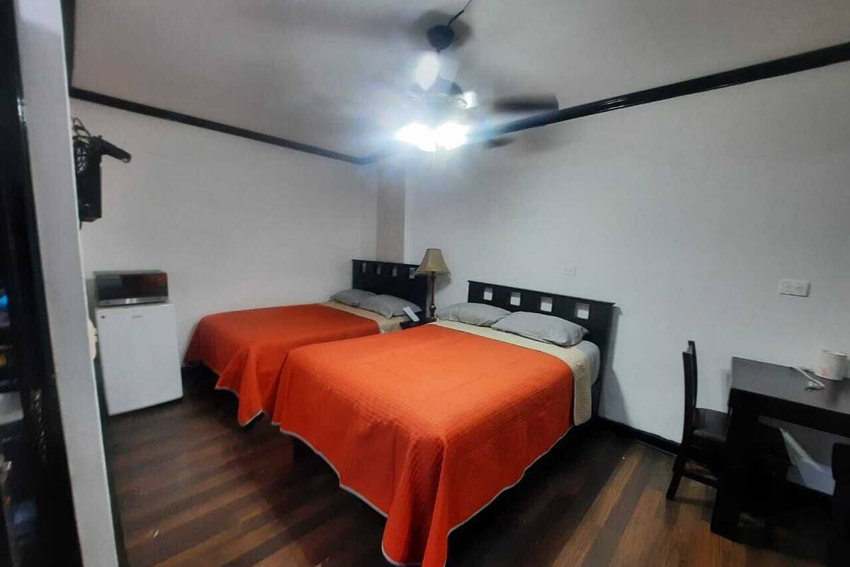 Eine kleine Einzelunterkunft mit zwei Betten, einem Deckenventilator, einem Schreibtisch und einer kleinen Küchenzeile.