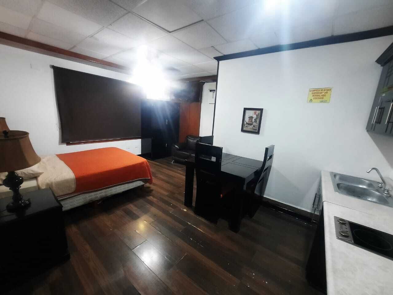 Una habitación pequeña con cama, escritorio, silla, una mesita y una zona de kitchenette.