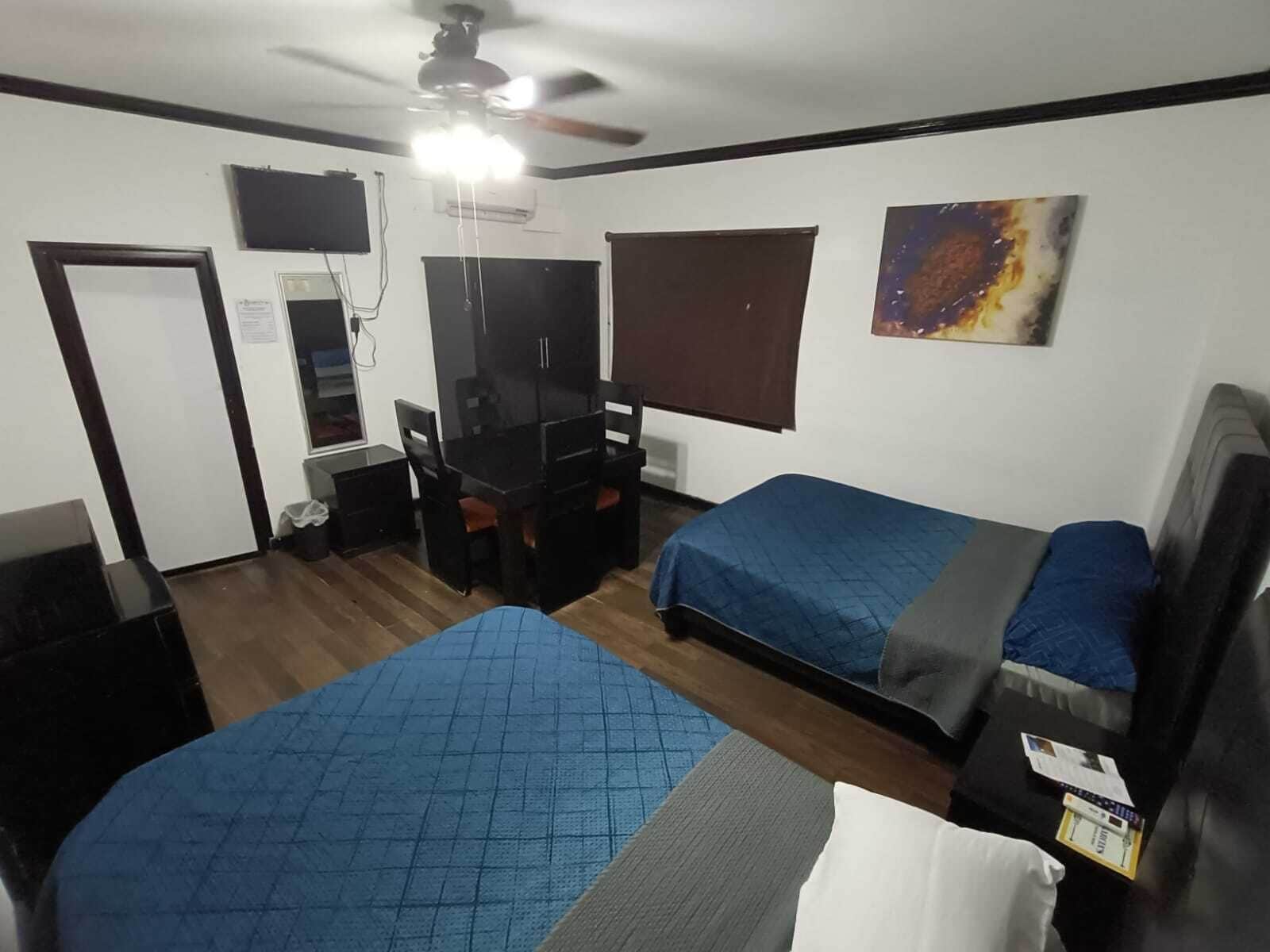 Una habitación con dos camas, un escritorio, una silla, un ventilador de techo y una ventana con persianas.