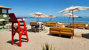 Un área de estar frente a la playa con sillas amarillas y rojas, sombrillas blancas y una silla de salvavidas.