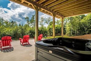 A covered outdoor area with a hot tub, red Adirondack chairs, and a view of a forest.