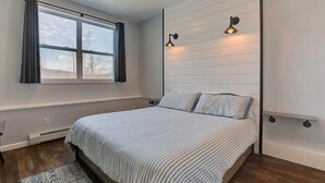 Une chambre à coucher avec un lit, une fenêtre avec des rideaux, un luminaire fixé au mur et un radiateur.