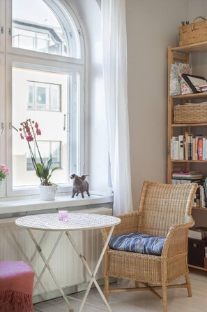 A room with a wicker chair, a small table, a bookshelf, and a window with curtains. - Cosy Quiet Apartment Close to the Sea and Parks (Helsinki)