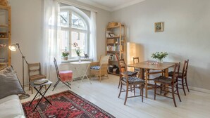 A cozy living room with a dining area, a bookshelf, and a large window with curtains. - Cosy Quiet Apartment Close to the Sea and Parks (Helsinki)