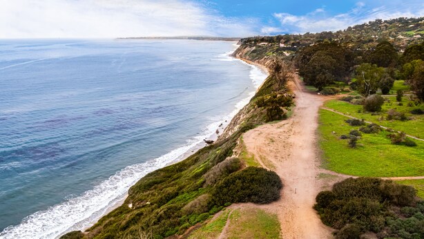Un paisaje costero con un sendero de tierra a lo largo de la playa, vegetación verde y una vista distante de un pueblo.