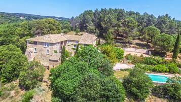 Une maison en pierre, avec un toit de tuiles, entourée d’une végétation luxuriante et surplombée d’un ciel bleu clair.