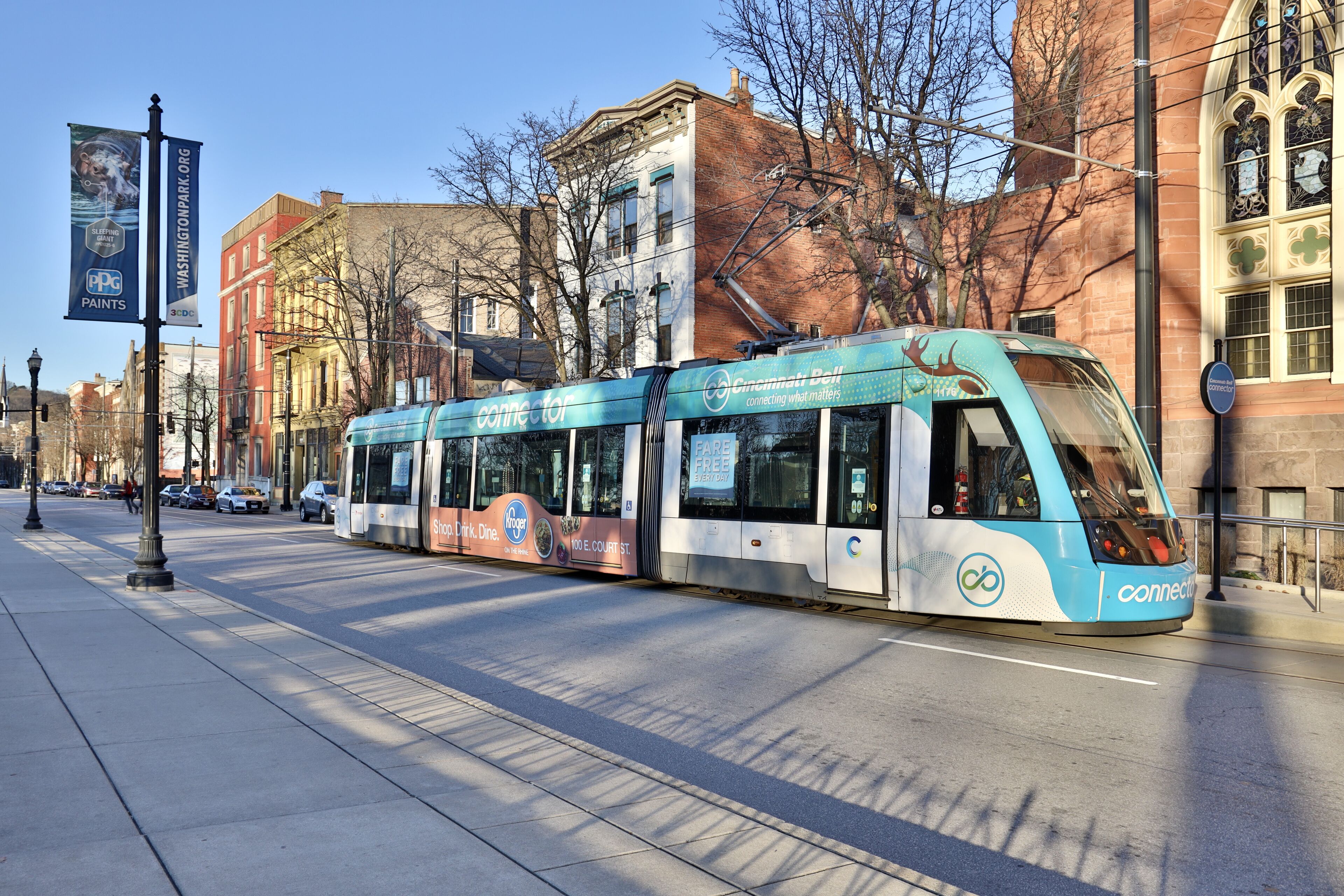 A modern tram with 'connect' branding, traveling on a city street with buildings and trees in the background.