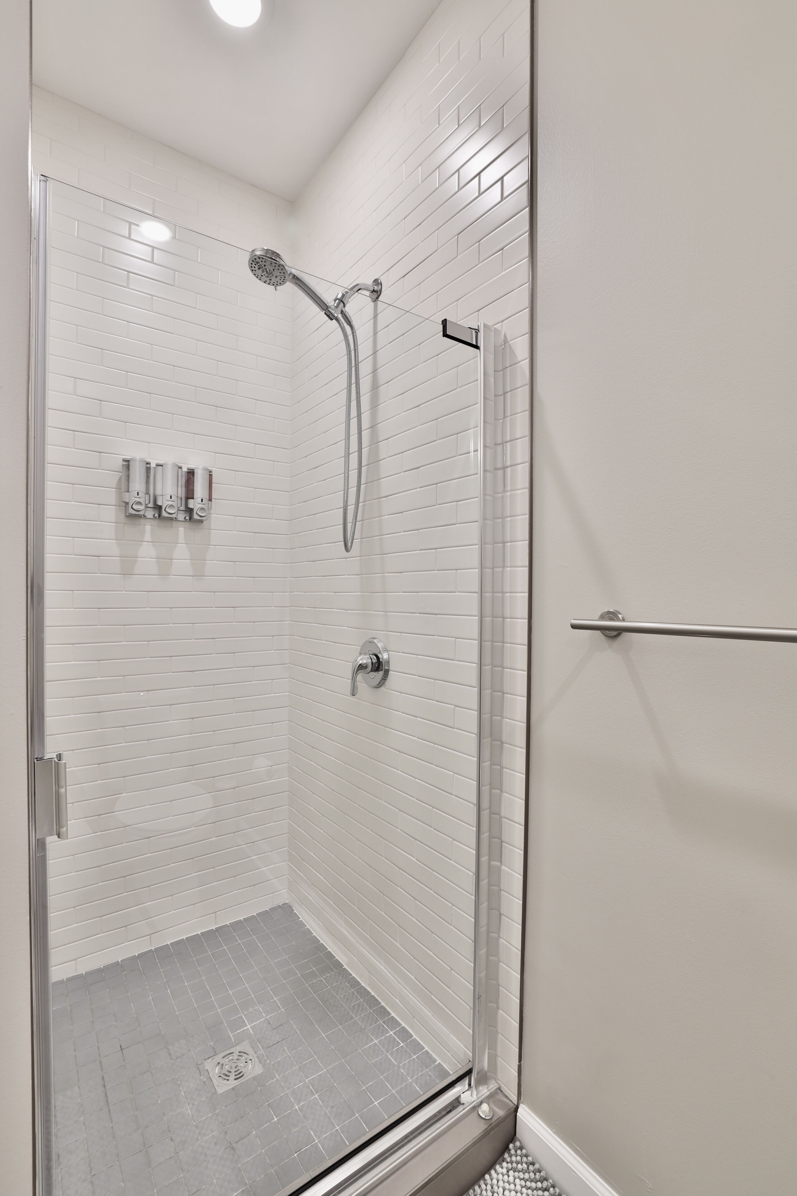 Une douche fermée par des parois de verre, avec des murs recouverts de carreaux blancs et une pomme de douche en métal.