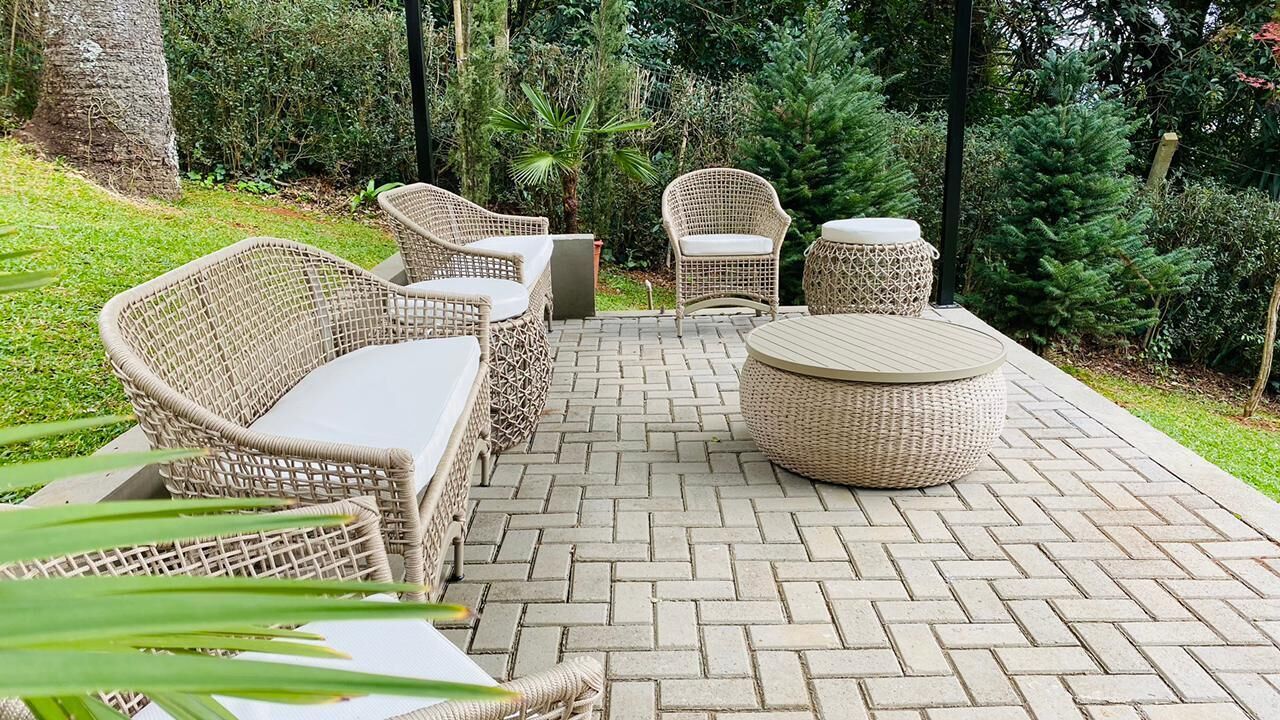 A patio with wicker furniture, including a sofa, chairs, and a round table, surrounded by a brick floor and greenery.