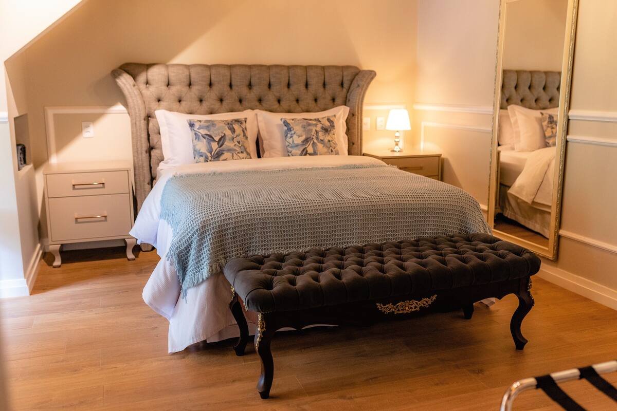A bedroom with a tufted headboard, a bed with patterned pillows, a bedside table with a lamp, and a large mirror on the wall.
