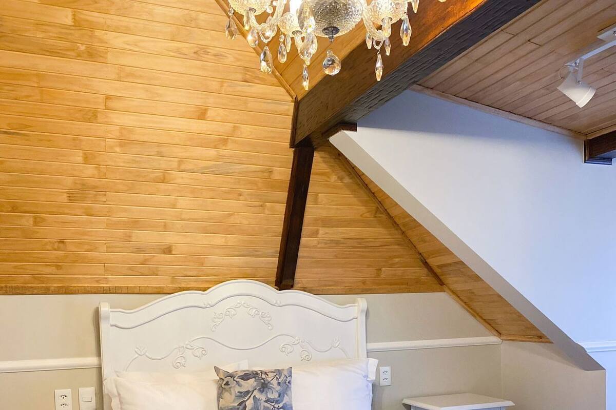A bedroom with a wooden ceiling, a chandelier, a bed with white bedding, a nightstand, and a small table.
