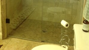 Un box de ducha con paredes de vidrio, una barra de apoyo, un inodoro con dispensador de toallas de papel y una botella a la derecha.