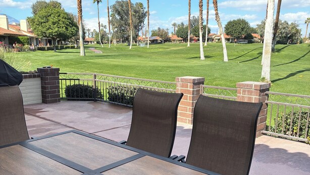 Un patio al aire libre con mesa y sillas, un muro de ladrillo y vistas a un campo de golf con palmeras.