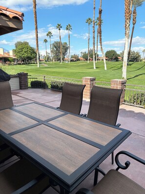 Une terrasse extérieure avec une table et des chaises, un mur en briques et une vue sur un terrain de golf bordé de palmiers.