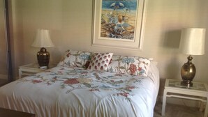 Una cama bien hecha con ropa de cama floral, dos mesitas de noche con lámparas y un cuadro con una playa en la pared.