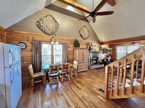 A cozy cabin interior with a dining area, wooden floors, a fireplace, and a ceiling fan.