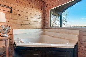 Interior de una cabaña de madera con una bañera grande, un ventanal con vistas a árboles y un ventilador sobre una mesa de madera.