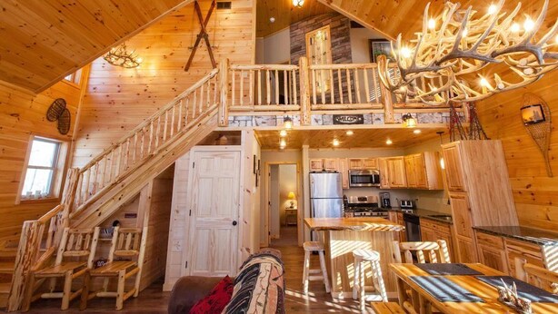 A cabin interior with a wooden staircase, a dining area, kitchen, and a chandelier made of antlers. - Beautiful ADK Getaway! Trail 4 and Lake Access!  Weekly Discount Available (Old Forge)