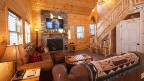 A cozy log cabin interior with a fireplace, a television mounted above it, a staircase, and a comfortable seating area with a sofa and a coffee table. - Beautiful ADK Getaway! Trail 4 and Lake Access!  Weekly Discount Available (Old Forge)