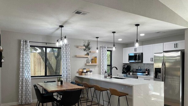 A modern kitchen with a central island, stainless steel appliances, pendant lighting, and a dining area with bar stools. - Great location! All New Super Private Modern Freshly Remodeled Retreat! (Cedar Park)