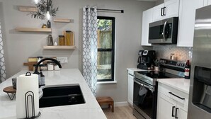 A modern kitchen with white cabinets, a stainless steel refrigerator, a built-in oven, and a microwave. There is a window with a view of greenery outside, and a wooden shelf with kitchen items. - Great location! All New Super Private Modern Freshly Remodeled Retreat! (Cedar Park)