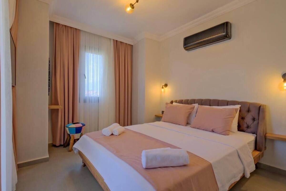 Quarto de hotel com uma cama grande, uma mesa de cabeceira, um pequeno banquinho e uma janela com cortinas.