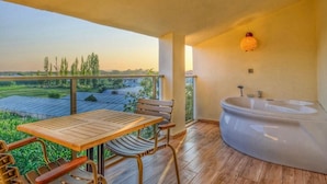 A balcony with a wooden table and chairs, a large bathtub, and a view of greenery and buildings.
