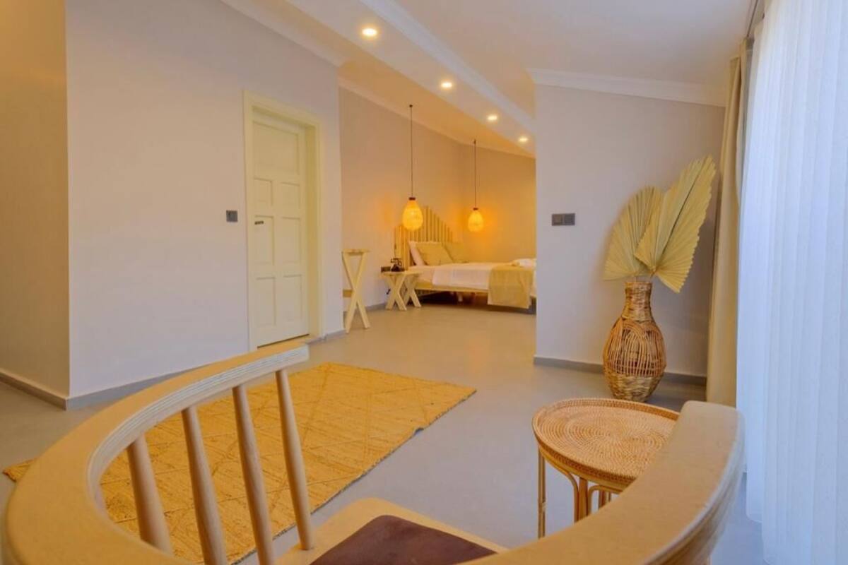Quarto de hotel com cama, uma mesinha, uma cadeira, um vaso grande e uma janela com cortinas.
