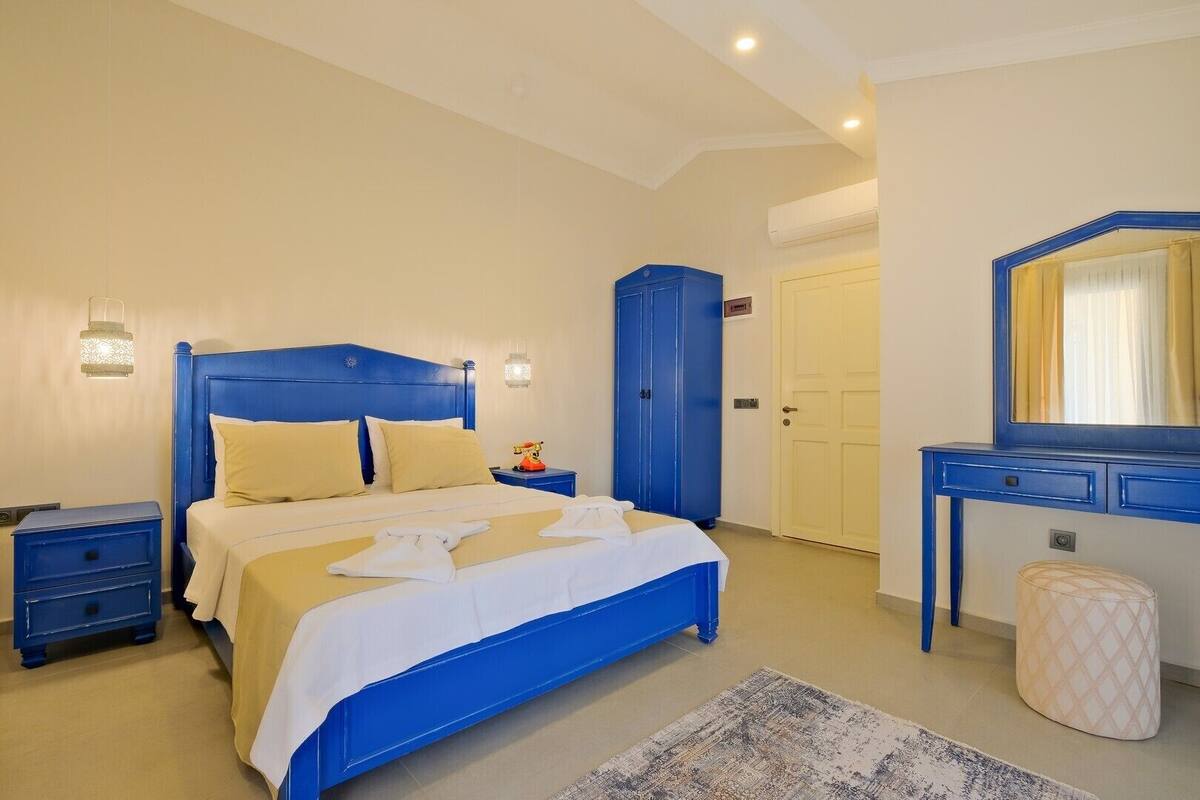 Quarto com uma cama azul, mesa de cabeceira, guarda-roupa, espelho e um pequeno banquinho.