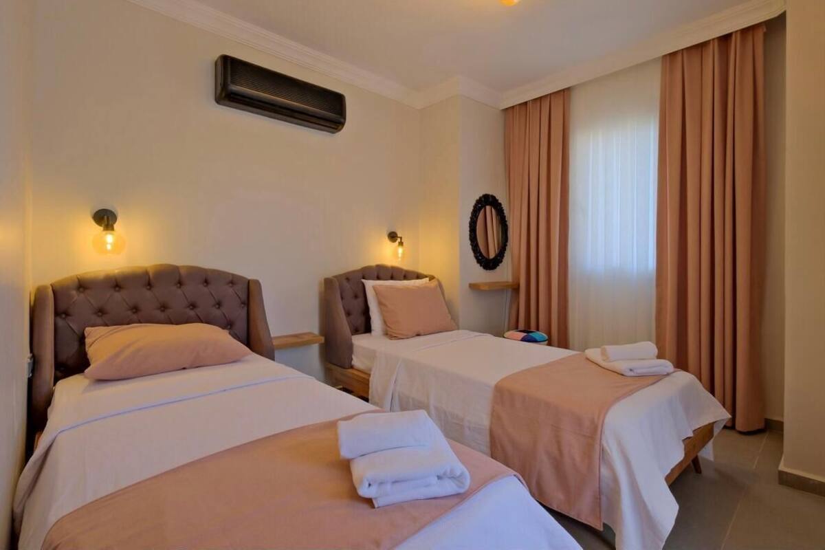 Quarto de hotel com duas camas, cada uma com cabeceira e almofadas, ar condicionado fixado na parede, um espelho e uma janela com cortinas.