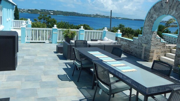 Patio esterno con vista sul mare, con un tavolo lungo e sedie, una pianta in vaso e un muro in pietra.