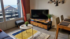 A living room with a TV, a small table with a plant, a sofa, a dining table, and a window with a view of buildings.