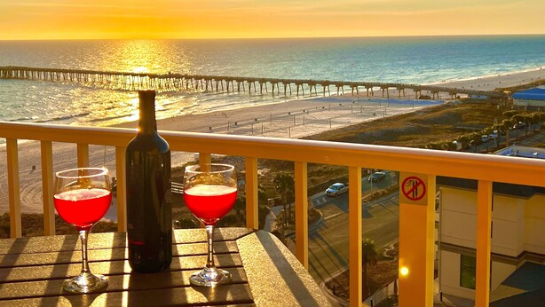 A balcony with a sunset view, a bottle, and two glasses of red drink. - FALL DEALS! CALYPSO 3-1004 FREE BEACHSETUPS! Romantic Ocean View from KING Bed! (Panama City Beach)