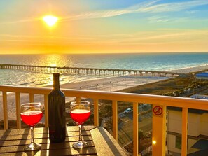 Un balcon avec vue sur le coucher de soleil, une bouteille et deux verres d’une boisson rouge.