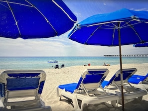 Ein Strand mit blauen Sonnenschirmen, weißen Liegestühlen und einem Steg im Hintergrund.
