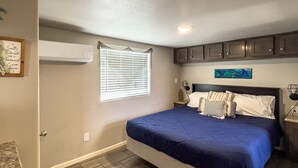 Un dormitorio con cama, ventana con persianas, aire acondicionado instalado en la pared y una mesita de noche con lámpara.
