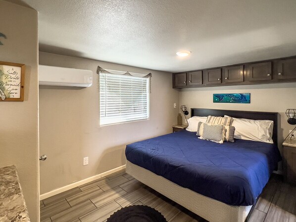 A bedroom with a bed, a window with blinds, a wall-mounted air conditioner, and a small bedside table with a lamp. - Siren's Adventure Riverfront Cottage! River Lodge NB31 (Parker Dam)