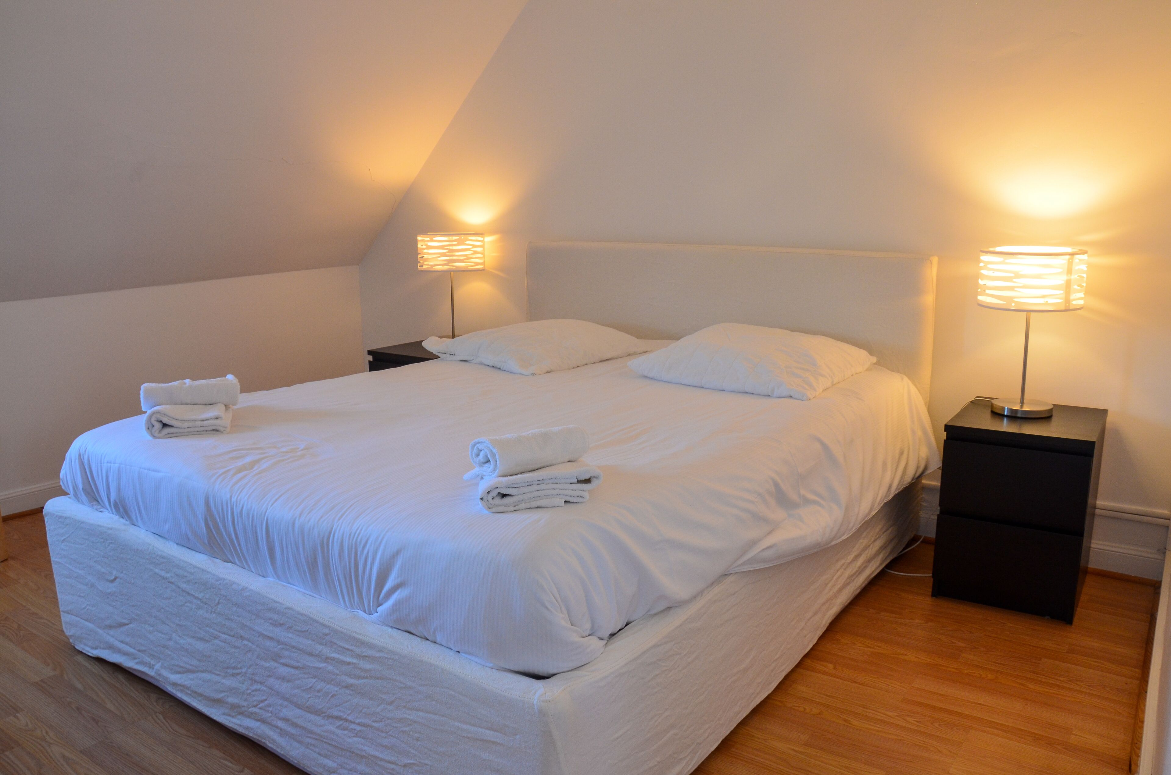 Een net opgemaakt bed met witte lakens en handdoeken, twee bedlampen en een houten vloer.