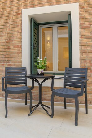 A table with two chairs outside a building with a brick wall and a window with green shutters. - Uni Island City Suites (Heraklion)