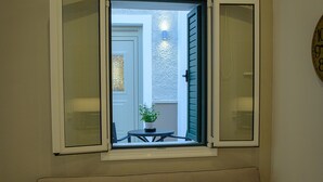 A living room with a beige sofa, a small round table, and a window with open shutters. - Uni Island City Suites (Heraklion)
