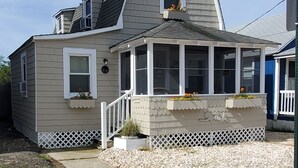 A two-story house with a gabled roof, a covered porch with white railings, and a small garden area with potted plants.